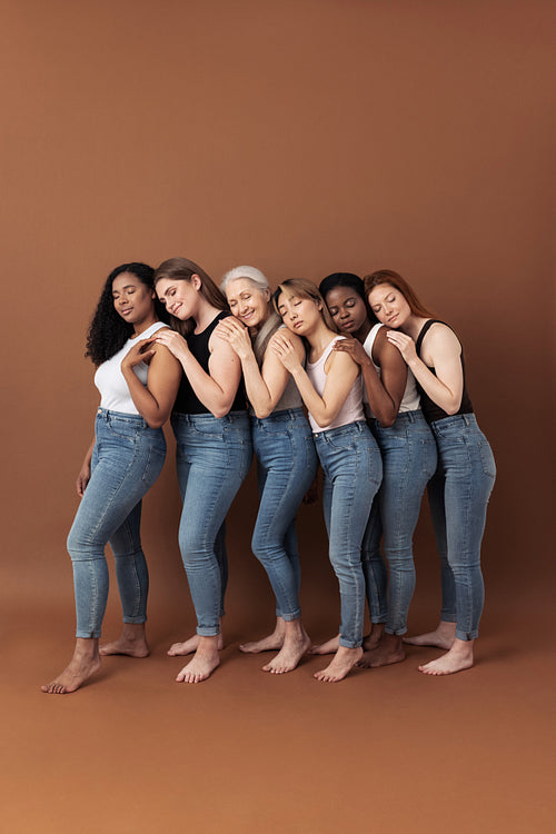 Six multiracial females in casuals hugging together on brown background in studio. Group of women of different ages standing together.
