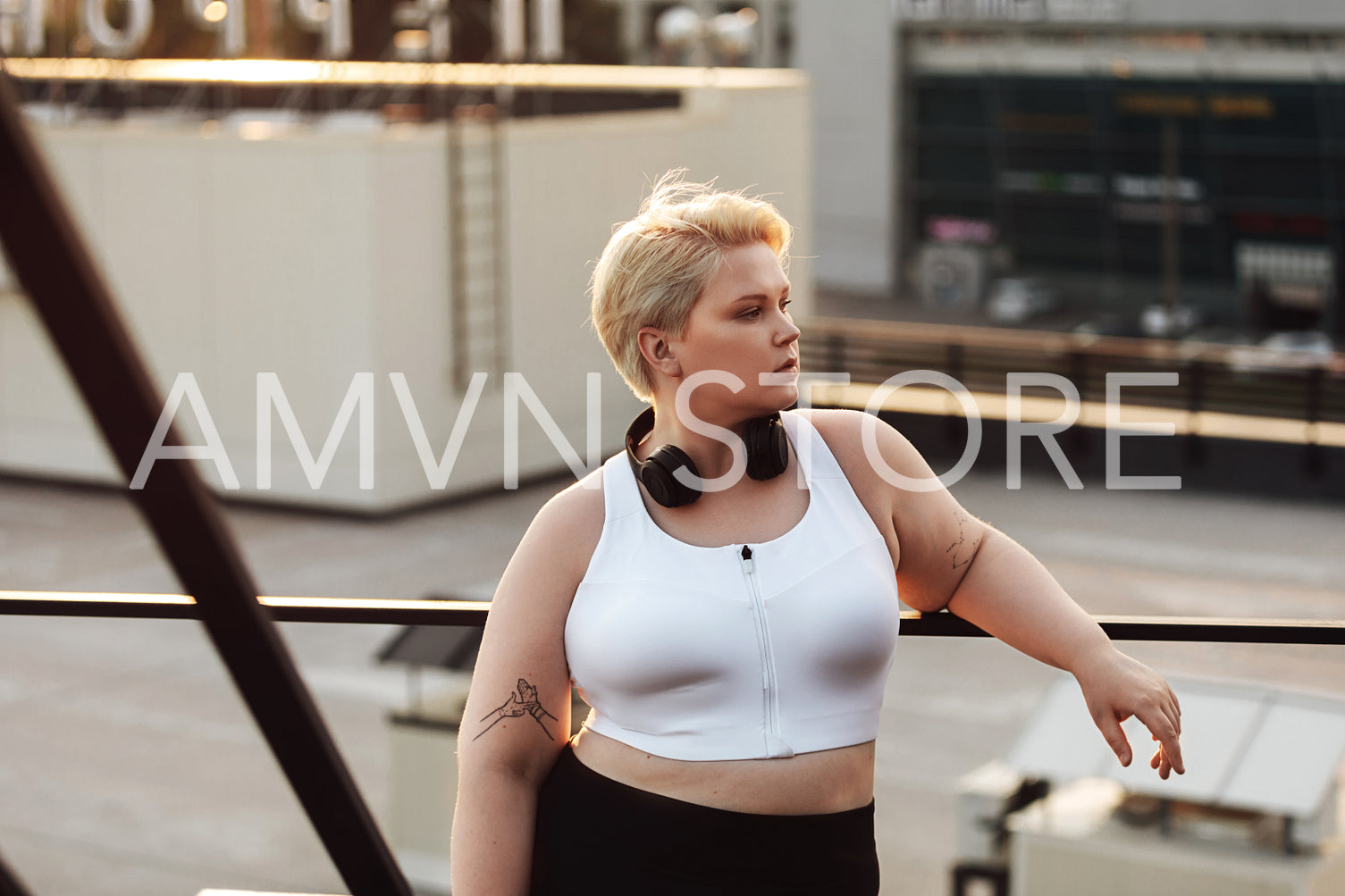 Woman with oversized body leaning to a railing during a workout on a roof. Oversize fitness woman in sportswear wearing headphones looking away outdoors.	