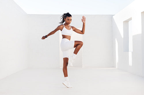 Young woman athlete jumping in white outdoor studio. Slim female doing cardio exercises. Sportswoman practicing high-intensity exercises.