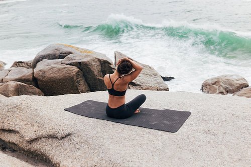 Rear view of young woman sitting on mat and warming up her neck by ocean. Female doing yoga and looking on waves.