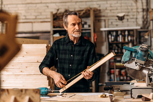 Mature carpenter measuring a length of wooden plank in workshop