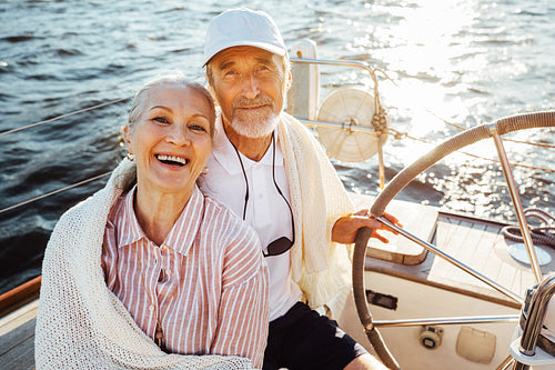 Senior couple sitting at the steering wheel on a sailboat and looking at camera