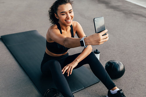 Female athlete sitting on a mat outdoors and taking selfie for social media. Smiling woman taking a break during training in the evening.