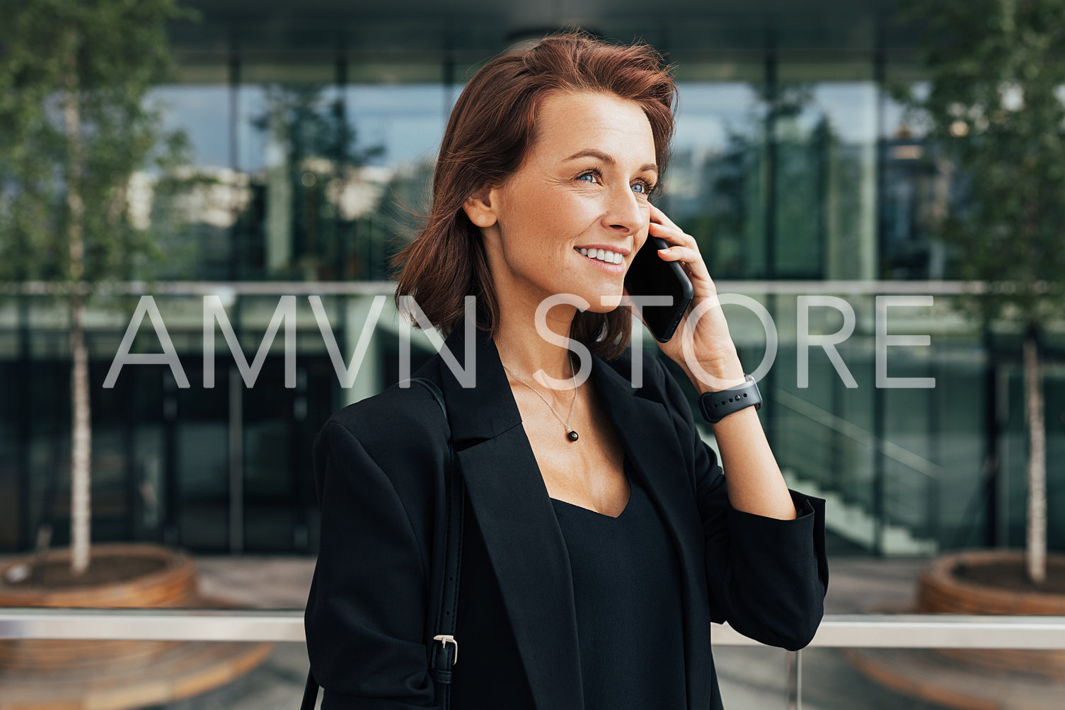 Smiling middle-aged woman talking on mobile phone at business building. Cheerful female with ginger hair in formal clothes standing outdoors and talking on her cell phone.