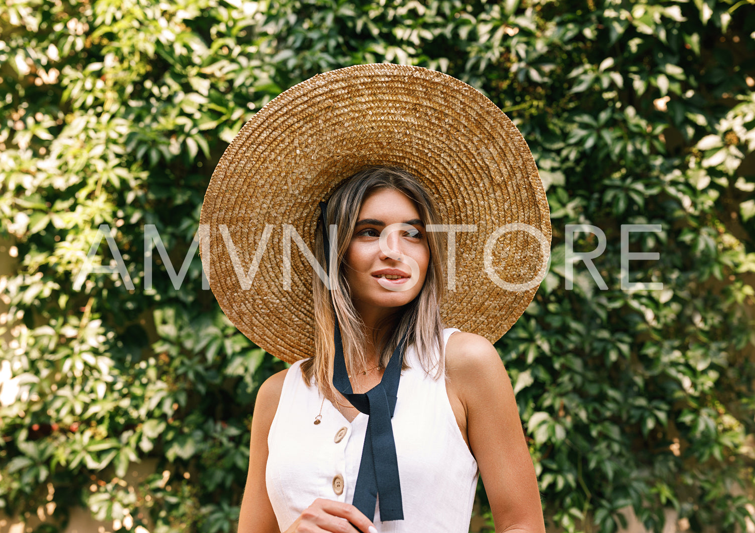 Blond woman in a straw hat standing outdoors in front of a wall with green leaves and looking away	