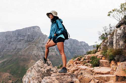 Woman wearing a hat and sports clothes relaxing during a mountain hike. Smiling female hiker looking down while standing on an edge.