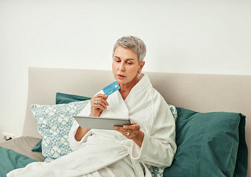 Senior woman shopping online while lying on a bed in hotel room