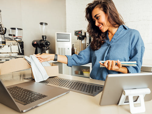 Young entrepreneur looking on pack of coffee and taking notes. Small cafe owner working at table with laptop and digital tablet.