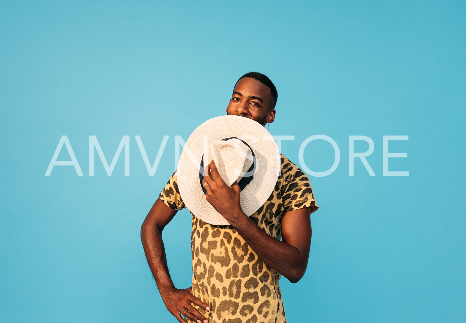 Happy man hiding his face with a straw hat and looking at camera against a blue background