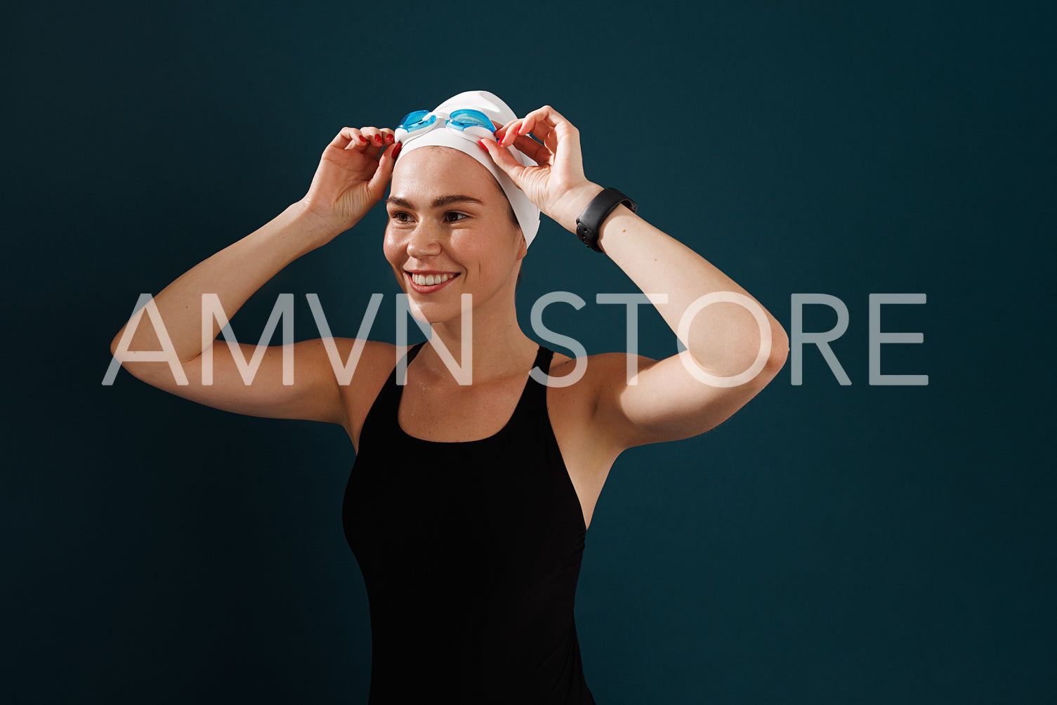 Smiling woman in swimwear adjusting her goggles while standing at blue backdrop