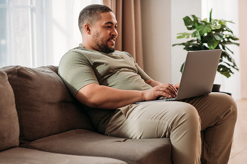 Smiling man with laptop on his hips sitting on a sofa. Young male working from home.