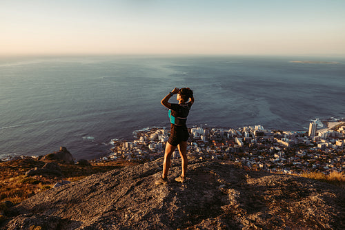 Woman in sportswear standing on the cliff and looking at the view at sunset. Female trail runner relaxing on mountain top looking at the ocean.