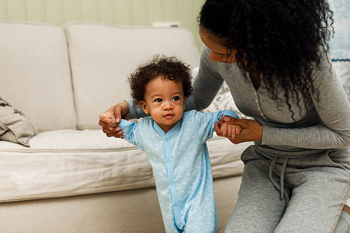 Mother sitting on the in a living room while helping her son walking