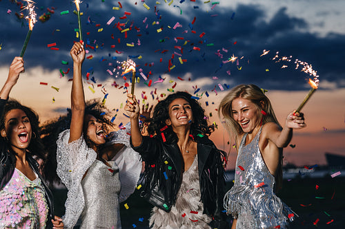 Group of four happy women dancing with sparklers under confetti at sunset