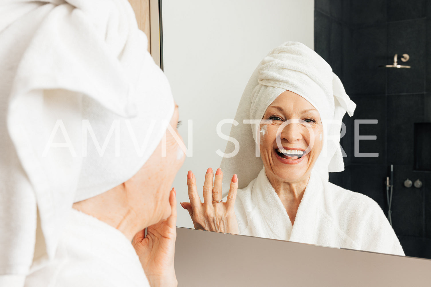 Laughing aged female with a wrapped towel on her head standing in front of a mirror in the bathroom