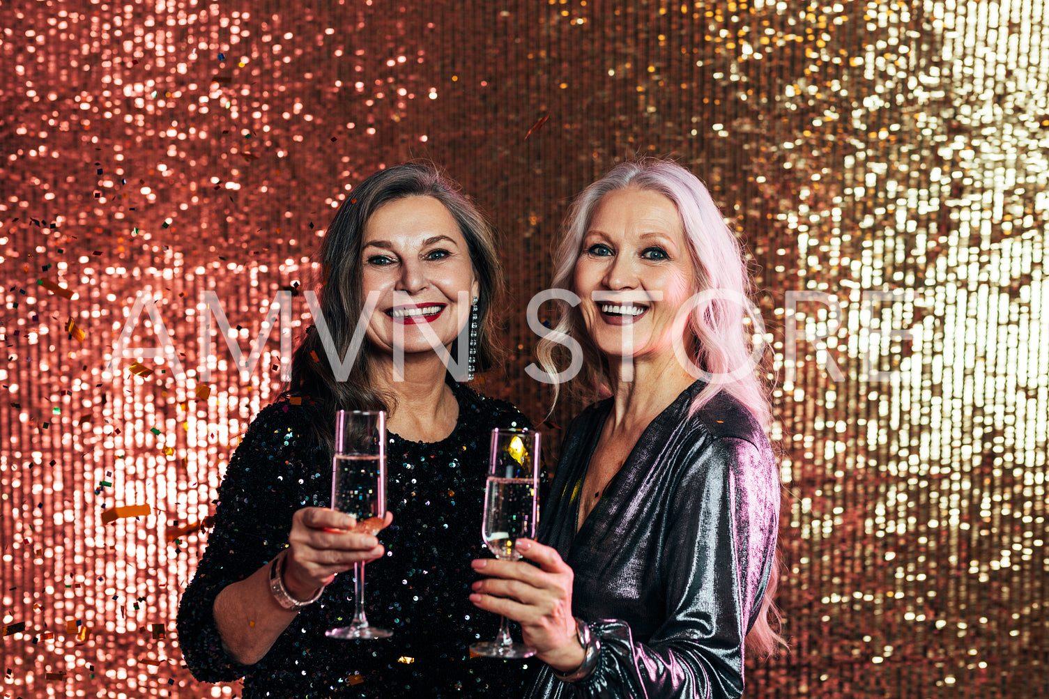 Stylish senior females standing together against glitter backdrop holding glasses of champagne