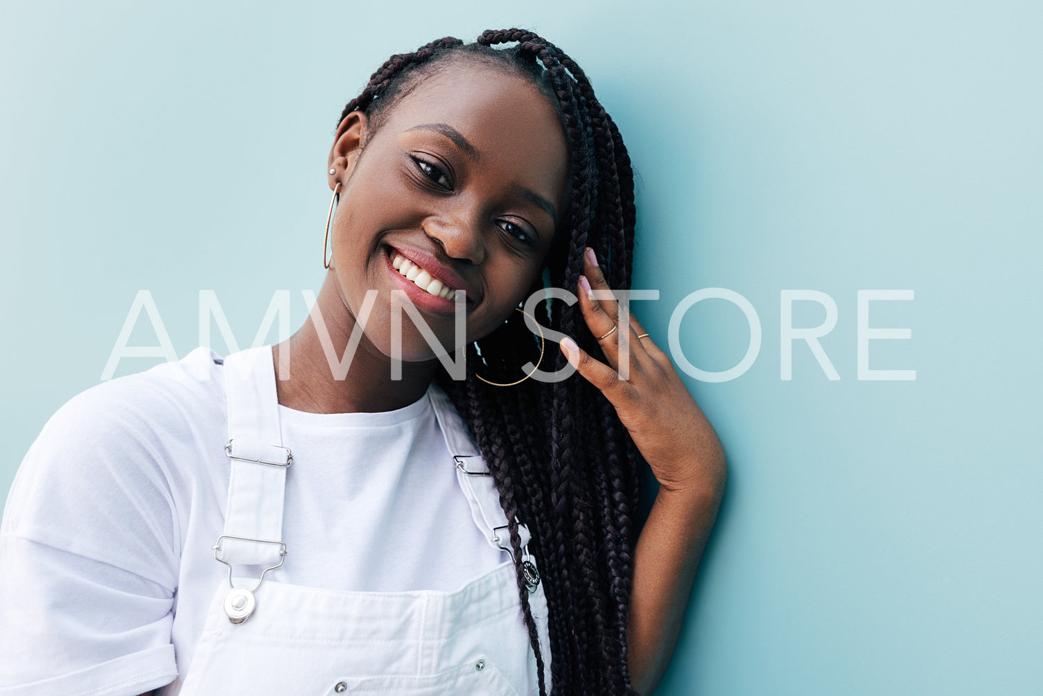 Close-up portrait of a young woman with braids. Smiling female in white clothes. Positive woman leaning blue wall and adjusting her braids while looking at camera.