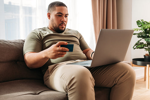 Young man paying online with a credit card at home. Mixed race male with a laptop on his hips making an internet order.