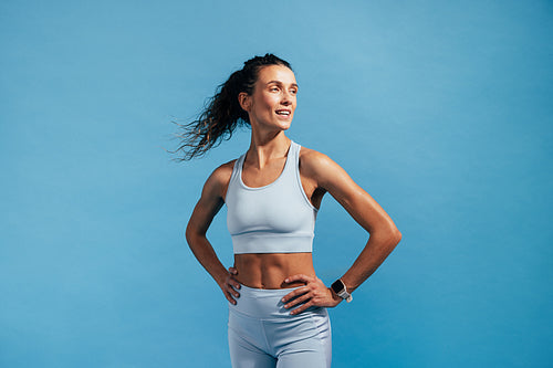 Smiling muscular woman in fitness wear standing on blue background. Female with hands on her hips relaxing after training.