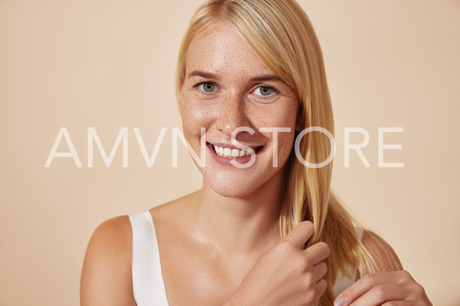 Beautiful smiling female adjusting her long blond hair while looking straight at a camera. Young woman with smooth skin posing in studio.