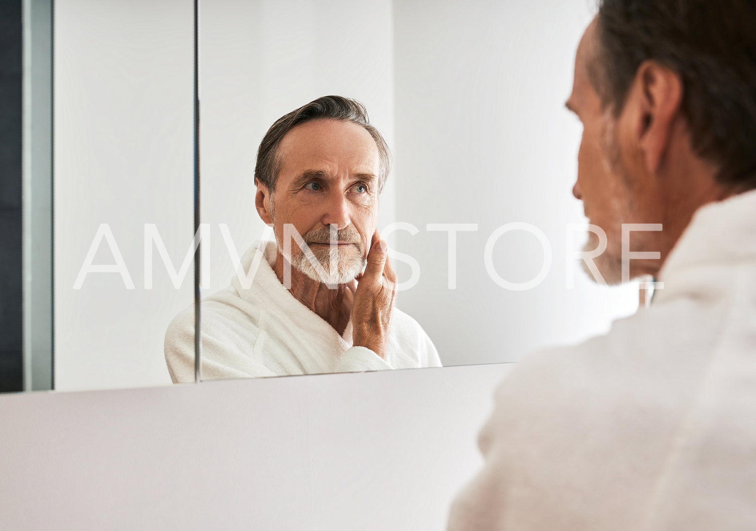 Mature man in a white bathrobe looking at a mirror in bathroom at morning	