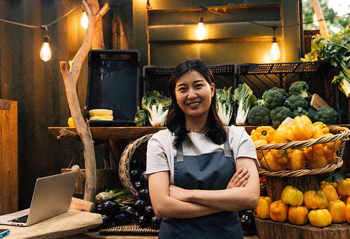 Portrait of an outdoor market owner. Asian woman with crossed arms wearing an apron standing against a stall.