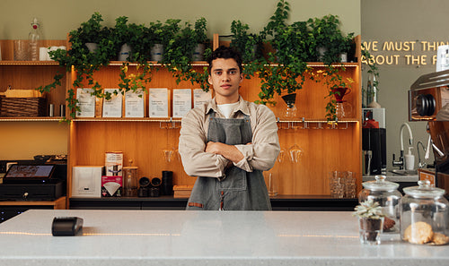 Young confident barista with cross hands looking straight at a camera while standing at a counter