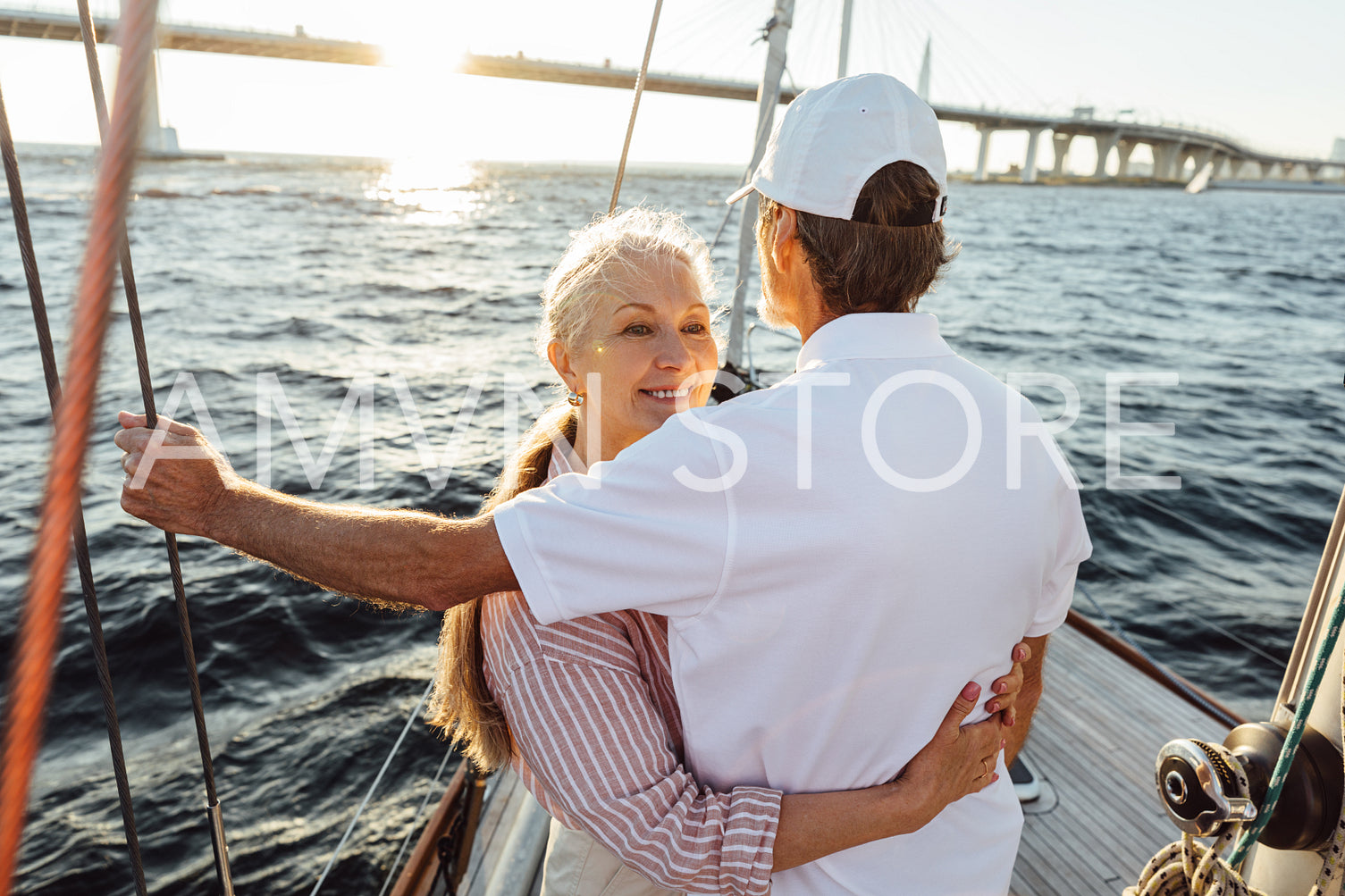 Smiling female looks over the shoulder of her husband and hugging him. Affectionate couple standing together on sailboat.	