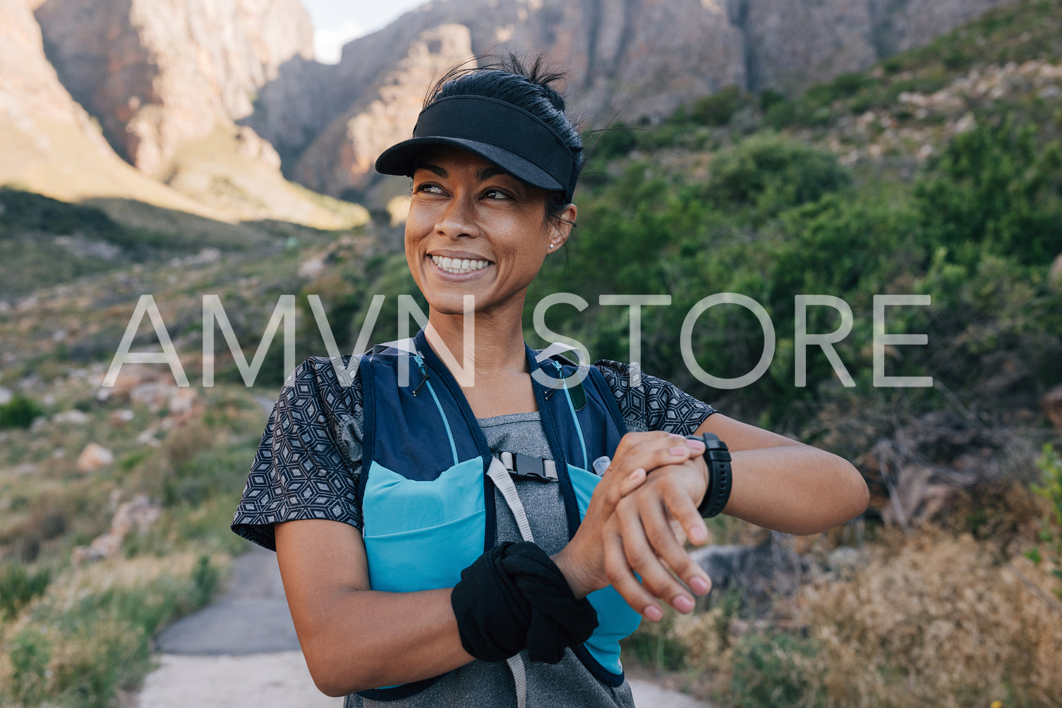 Smiling woman hiker in cap looking away. Female trail runner checking smart watch while standing in valley.