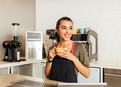 Smiling female barista looking away, holding a coffee mug in cafe