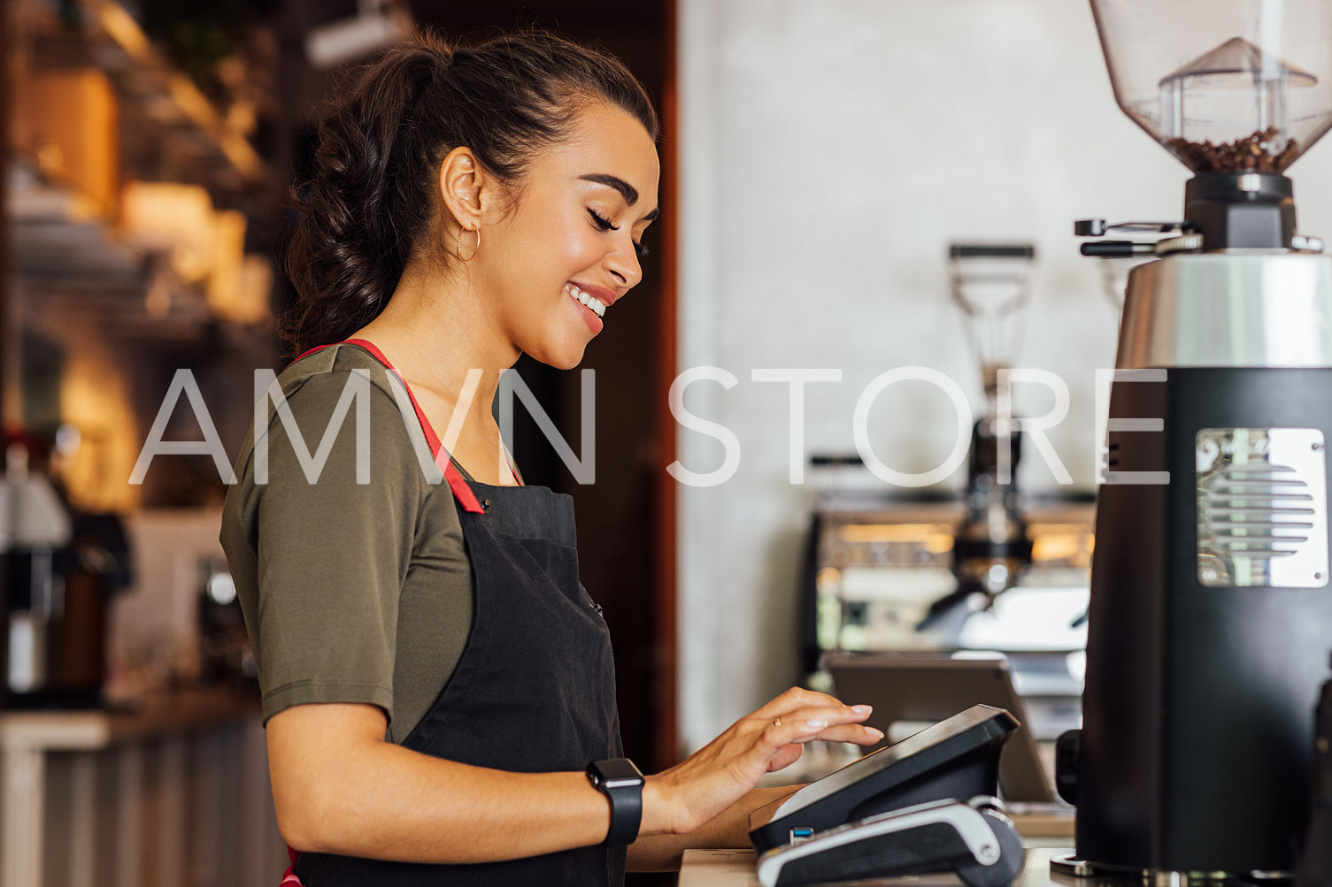 Side view of smiling waitress in apron typing digital cash terminal in coffee shop	