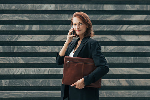 Smiling female with ginger hair talking on a mobile phone while standing outdoors. Middle-aged businesswoman in black clothes.