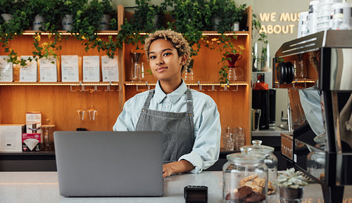 Young female coffee shop owner at counter with a laptop. Woman barista in an apron working in a coffee shop.