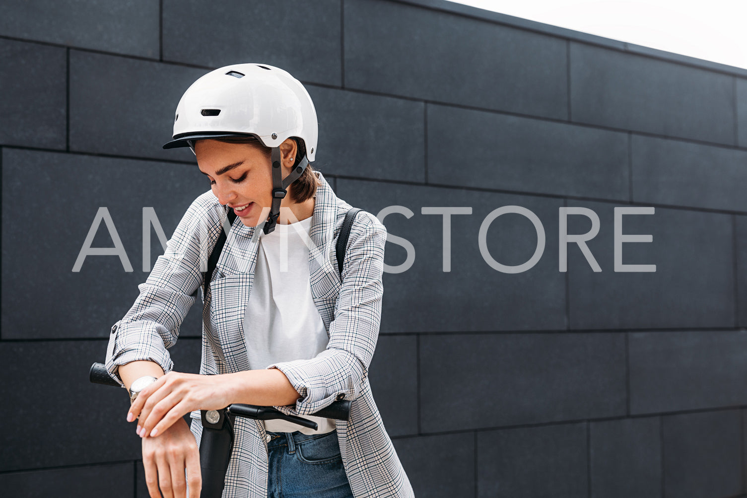 Woman in white helmet in formal wear standing at black wall outdoors leaning on electric push scooter