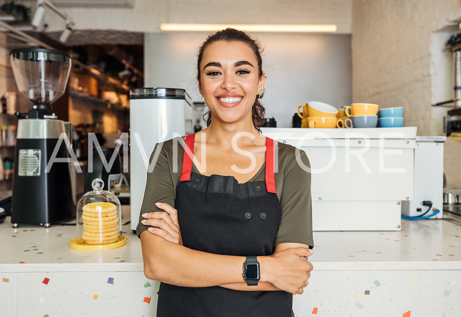 Female barista standing arms crossed in a cafe. Smiling waitress looking at camera while standing in front of a counter.