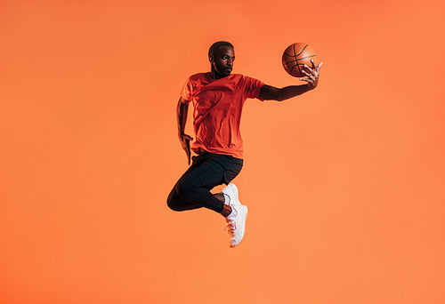 Young man in sportswear practicing basketball against an orange background. African male athlete jumping in the air with basket ball in studio.