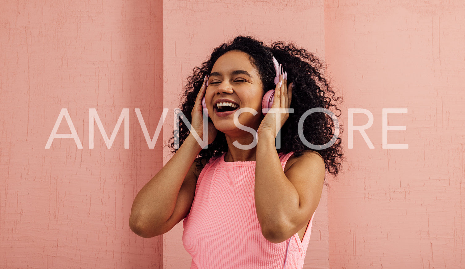 Laughing woman listening to music with pink headphones with closed eyes. Young happy female in pink casuals enjoying music.
