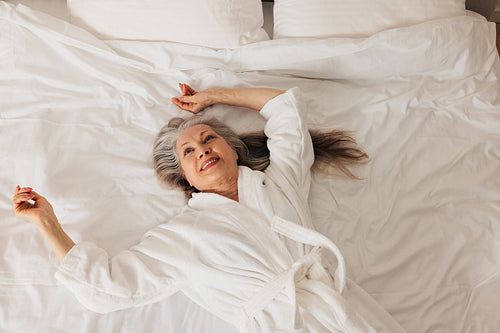 Senior woman in a bathrobe looking up while lying on a bed. Smiling retired female enjoying morning.