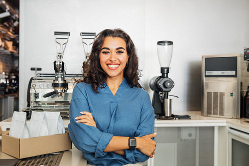 Smiling cafe owner looking at camera with arms crossed. Young female entrepreneur standing in a coffee shop at table.