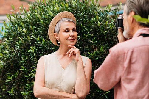 Smiling senior woman in a straw hat looking at the camera while her husband photographs her