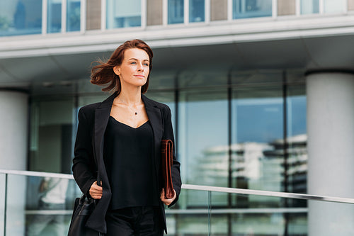 Confident middle-aged businesswoman holding a leather folder walking against an office building