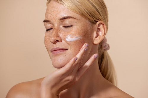 Close up of beautiful young female with cream on a face. Smiling woman with closed eyes touching cheek with fingers.