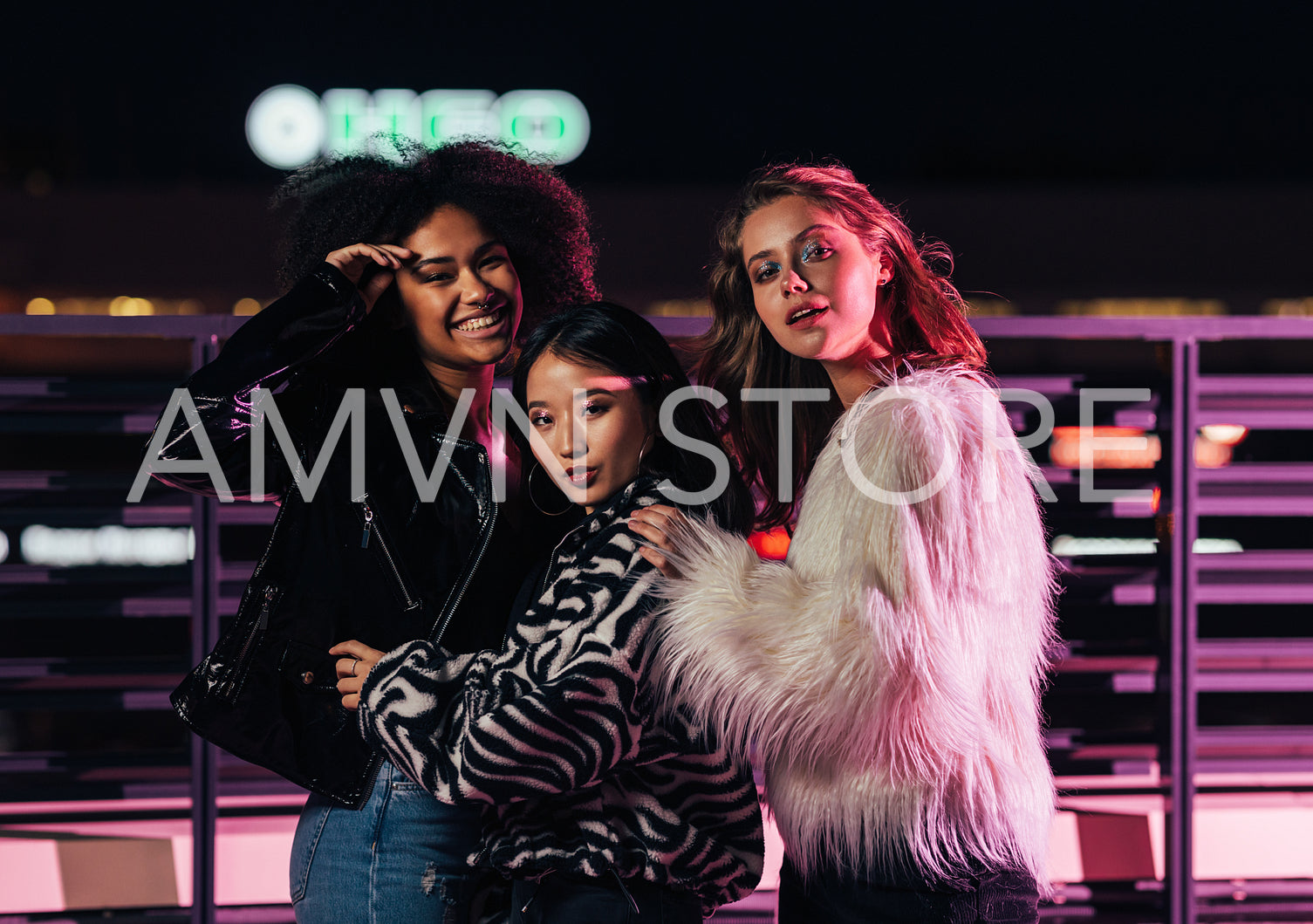 Three stylish girls standing on a street at night in front of neon lights and looking at camera	