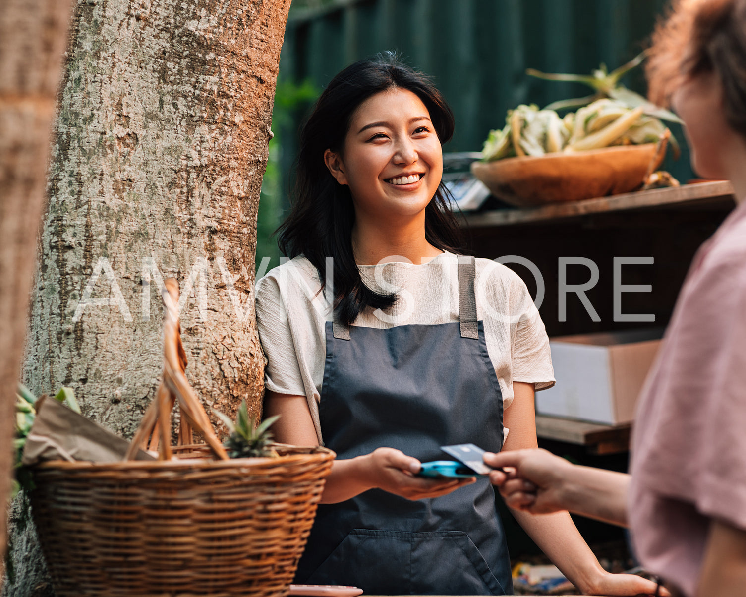 Happy Asian woman in an apron receiving payment on the outdoor market. Vendor with a card machine smiling and looking at the customer.