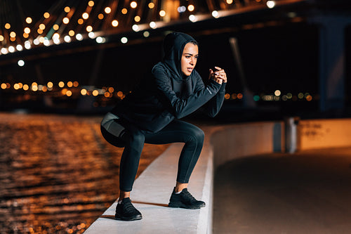 Woman in hooded sport shirt doing squats on embankment at night