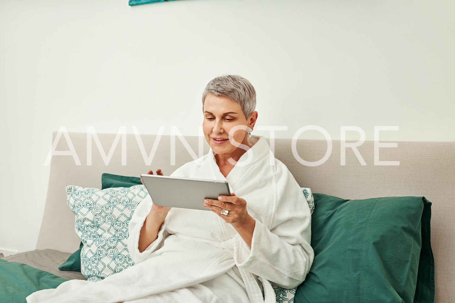 Smiling mature woman in bathrobe relaxing on a bed holding a digital tablet