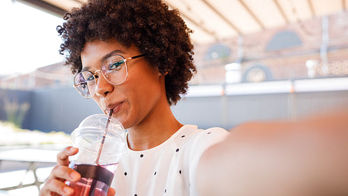 Close up of young woman drinking with straw while taking selfie on mobile phone, looking at frontal camera