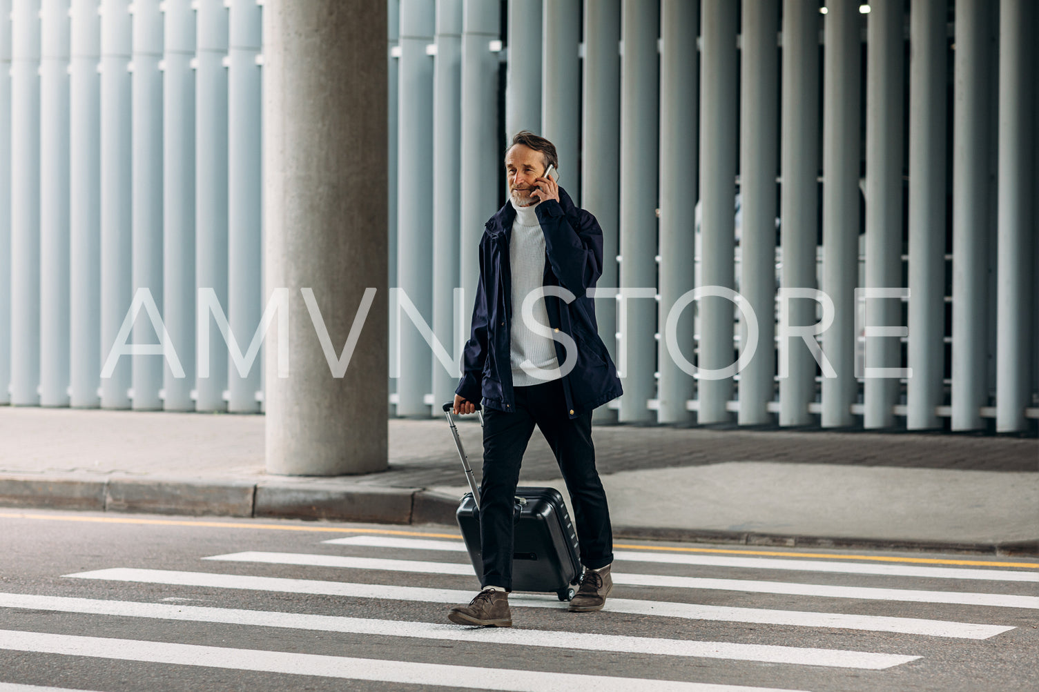 Senior man talking on cell phone while walking on pedestrian crosswalk and pulling suitcase	