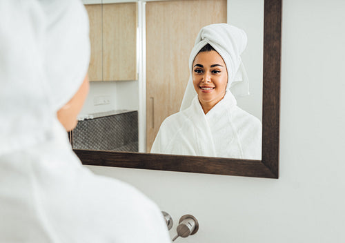 Woman in bathrobe looking at her reflection. Beautiful brunette with a white towel on her head.