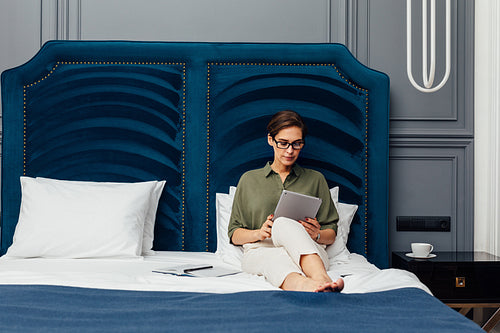 Woman entrepreneur lying on a bed in a luxury hotel room and reading from digital tablet